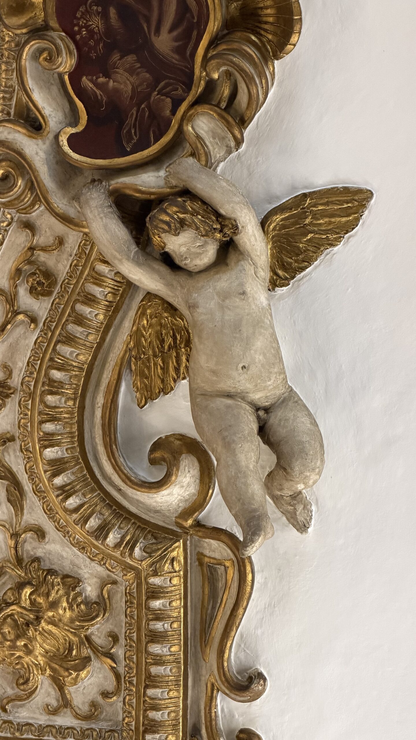 Putto alato in stucco con ali dorate, sospeso sulla cornice